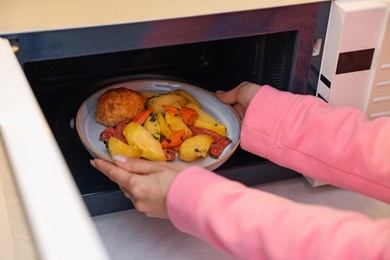 Woman putting plate with lunch into microwave indoors, closeup