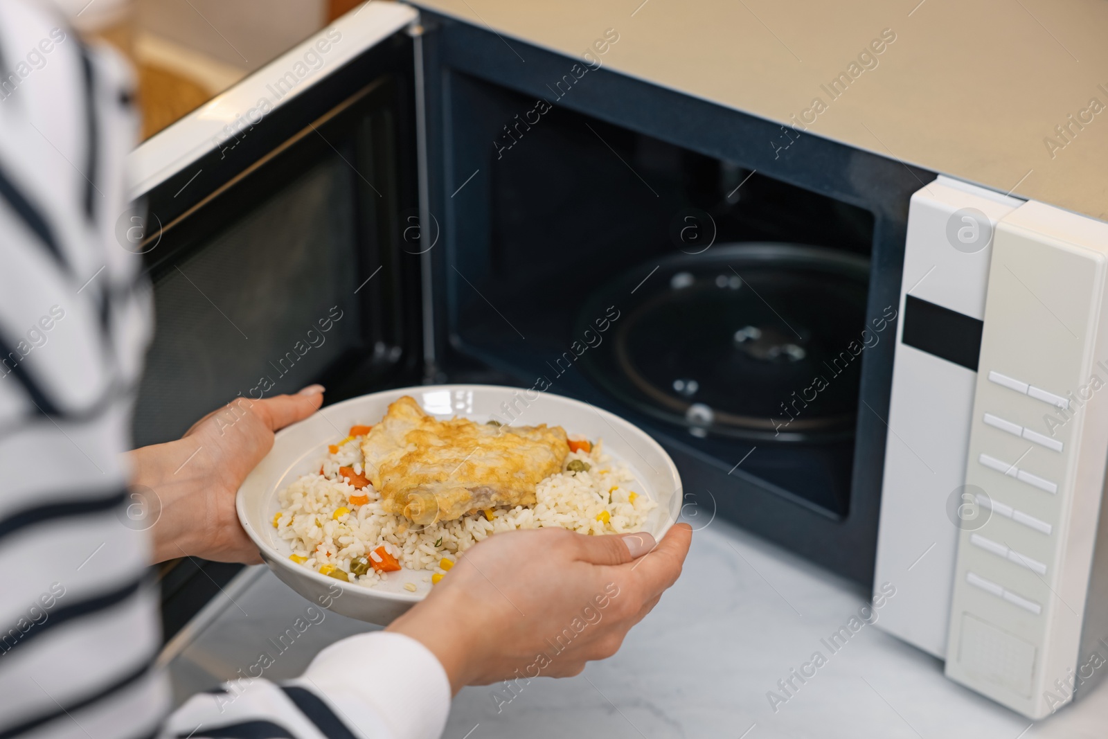 Photo of Woman putting plate with lunch into microwave indoors, closeup