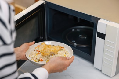 Photo of Woman putting plate with lunch into microwave indoors, closeup