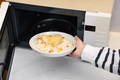 Photo of Woman putting plate with lunch into microwave indoors, closeup