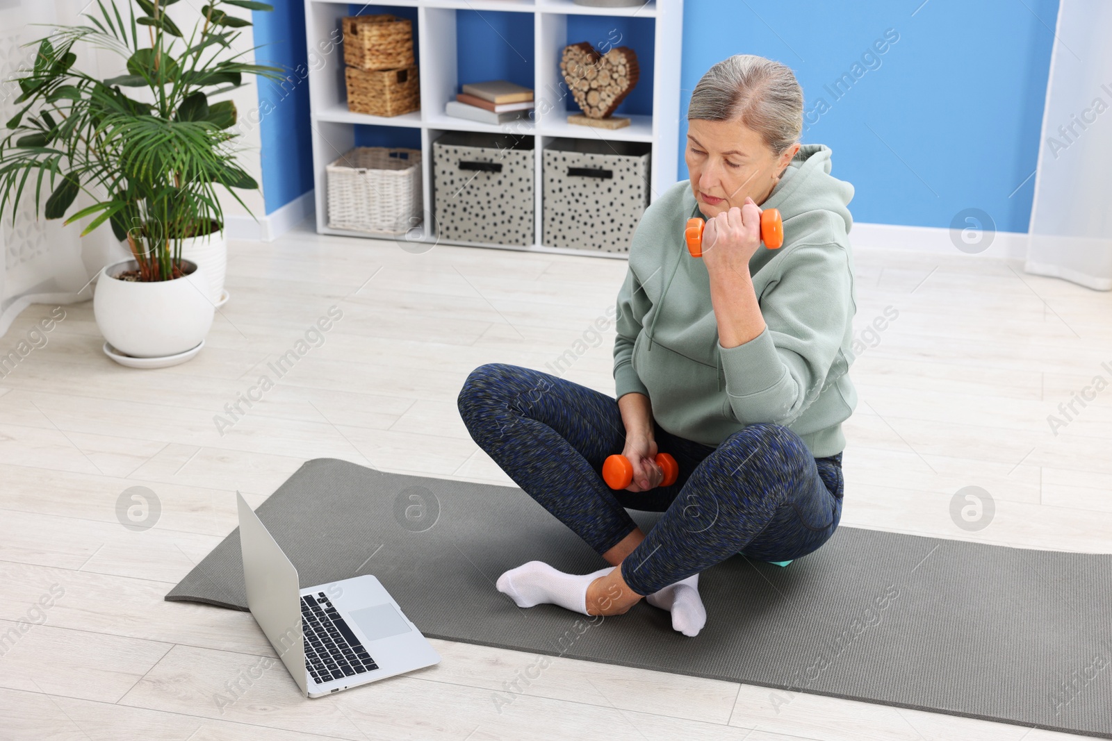 Photo of Elderly woman exercising with dumbbells near laptop at home