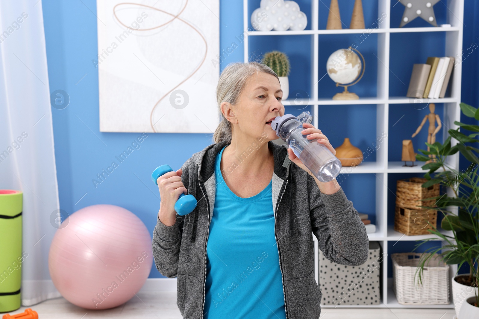 Photo of Elderly woman drinking water during exercise with dumbbell at home