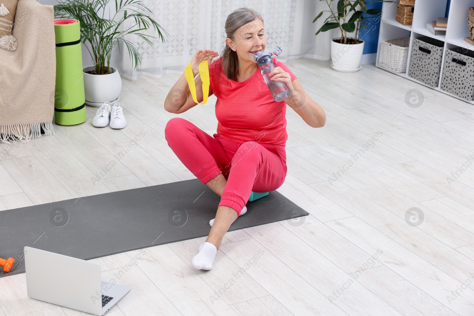 Photo of Elderly woman drinking water during exercise with fitness elastic band at home