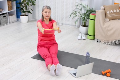 Elderly woman exercising with fitness elastic band near laptop at home