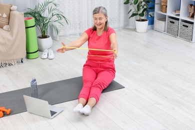 Photo of Smiling elderly woman exercising with fitness elastic band near laptop at home