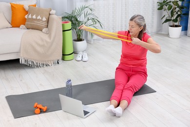 Elderly woman exercising with fitness elastic band near laptop at home