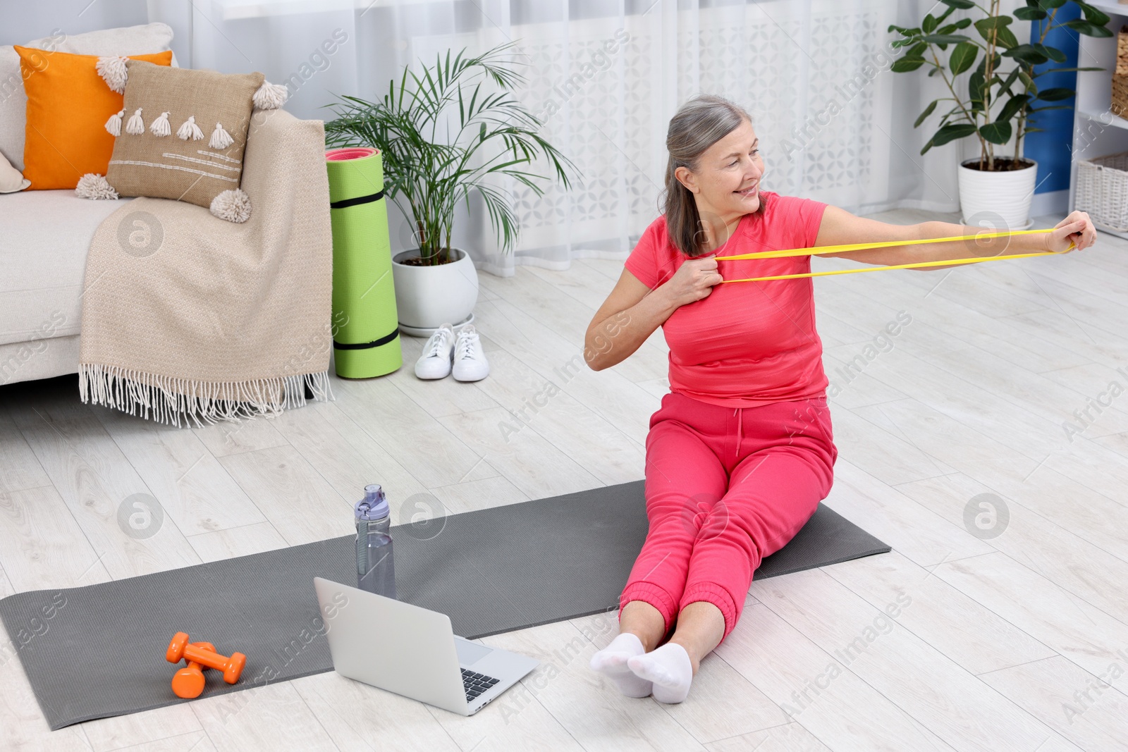 Photo of Smiling elderly woman exercising with fitness elastic band near laptop at home