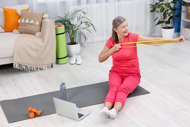 Smiling elderly woman exercising with fitness elastic band near laptop at home