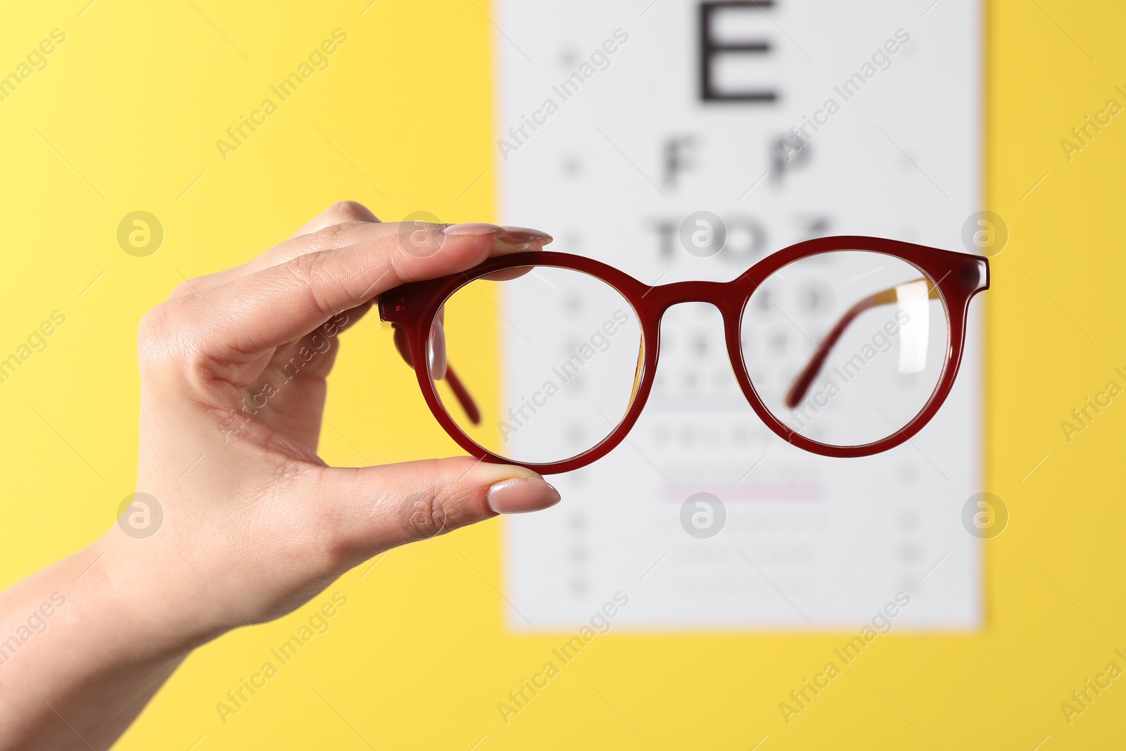 Photo of Woman holding glasses against vision test chart on yellow background, closeup