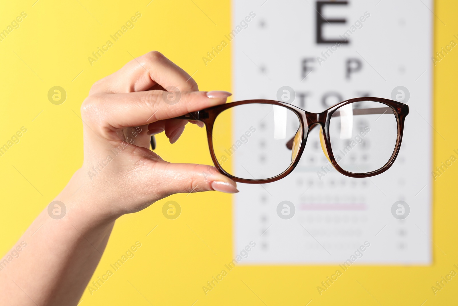 Photo of Woman holding glasses against vision test chart on yellow background, closeup