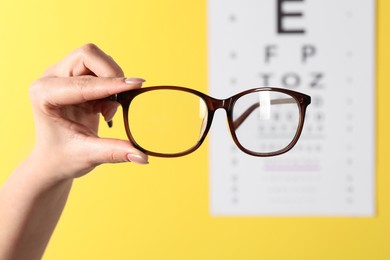 Photo of Woman holding glasses against vision test chart on yellow background, closeup