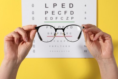 Photo of Woman holding glasses against vision test chart on yellow background, closeup