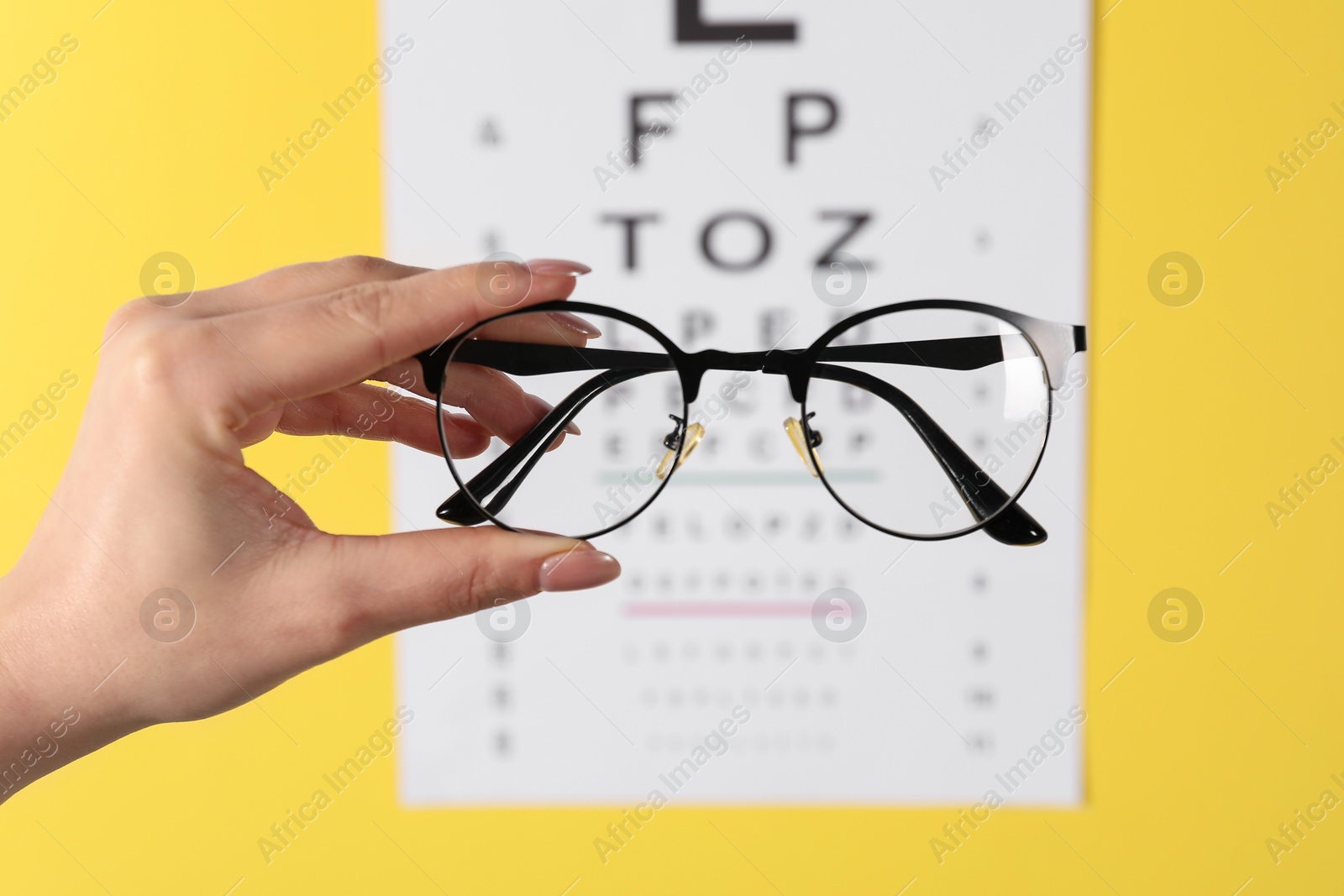 Photo of Woman holding glasses against vision test chart on yellow background, closeup