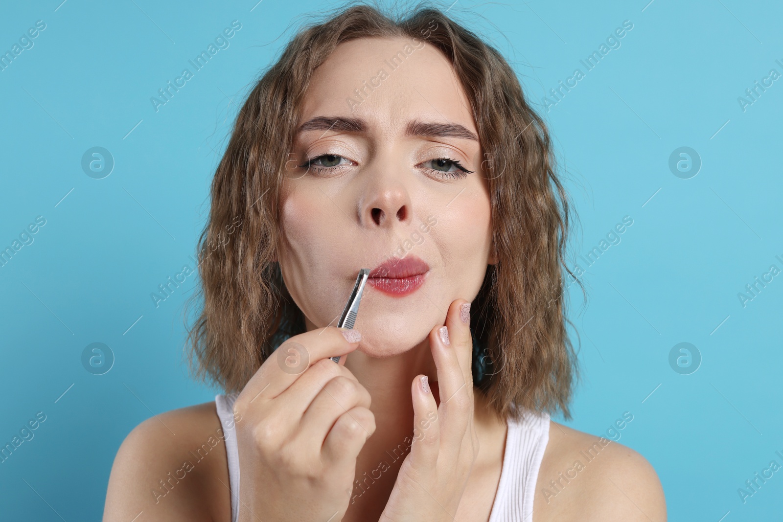 Photo of Beautiful woman plucking her mustache with tweezers on light blue background