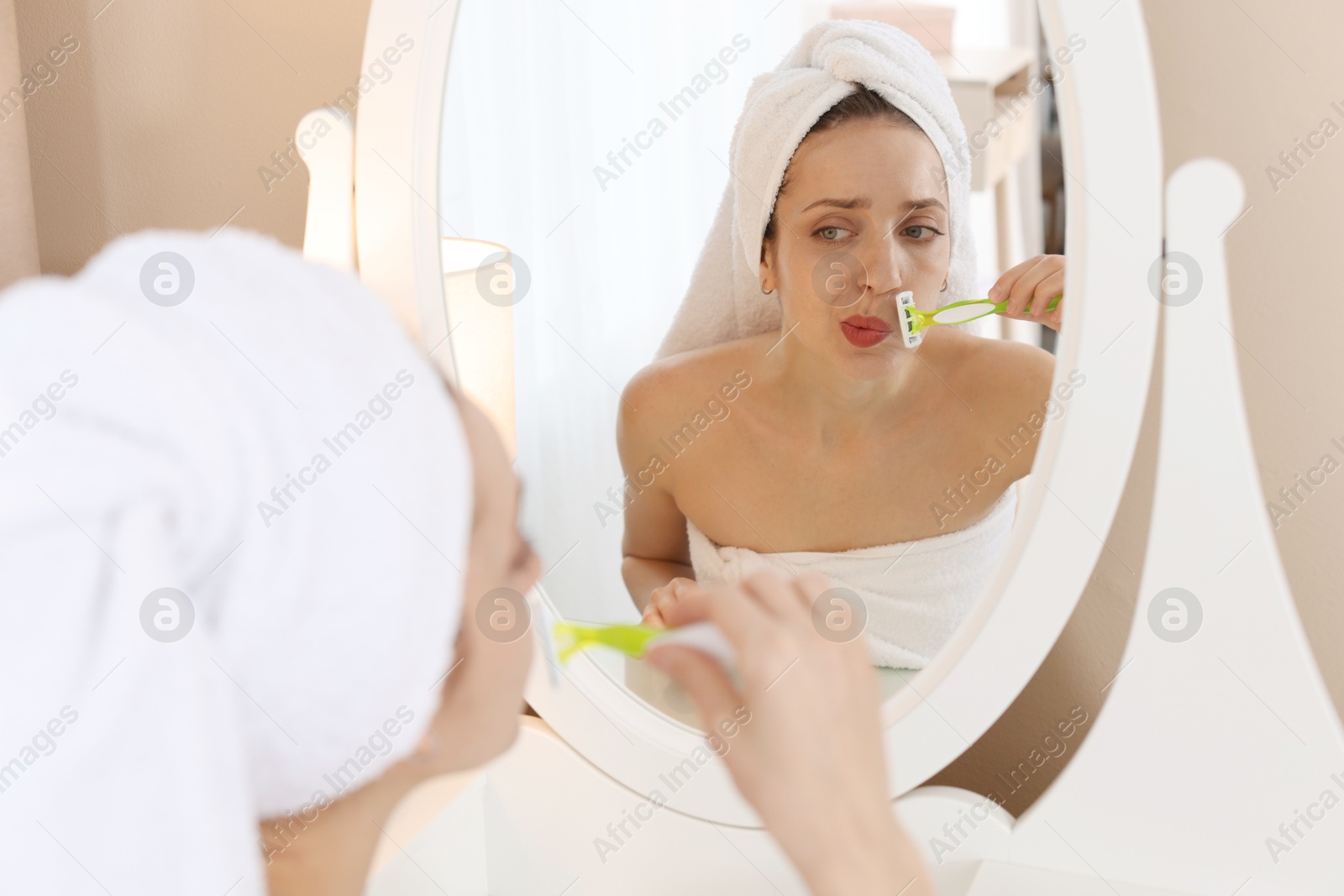 Photo of Beautiful woman shaving her mustache with razor near mirror at home