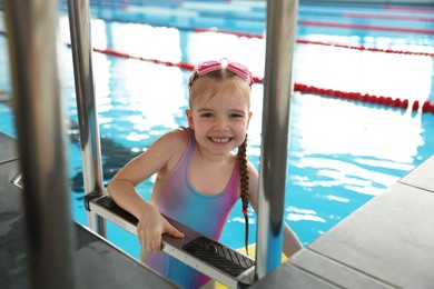 Little girl with goggles getting out of swimming pool indoors