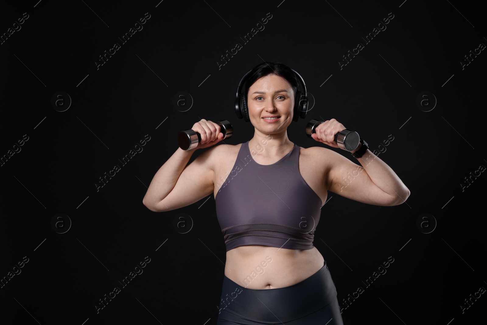 Photo of Plus size woman in gym clothes with headphones and dumbbells on black background