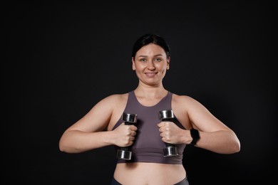 Plus size woman in gym clothes doing exercise with dumbbells on black background