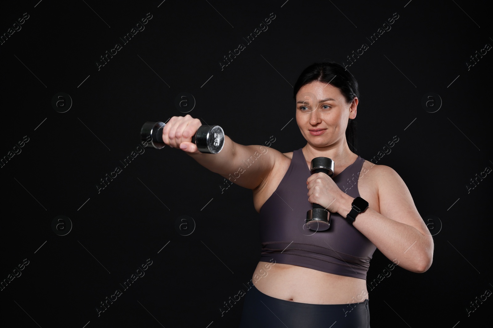 Photo of Plus size woman in gym clothes doing exercise with dumbbells on black background, space for text