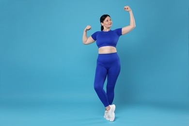 Plus size woman in gym clothes showing her biceps on light blue background, space for text