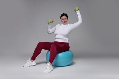 Plus size woman in gym clothes with dumbbells and fitness ball on grey background