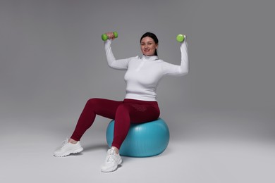 Plus size woman in gym clothes with dumbbells and fitness ball on grey background