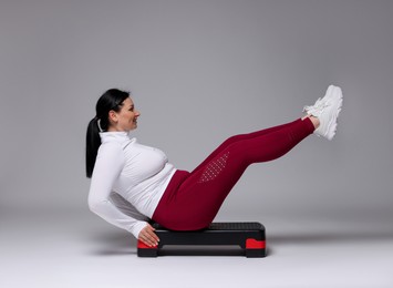 Photo of Plus size woman in gym clothes doing exercise with step platform on grey background