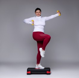 Photo of Plus size woman in gym clothes doing exercise with dumbbells and step platform on grey background