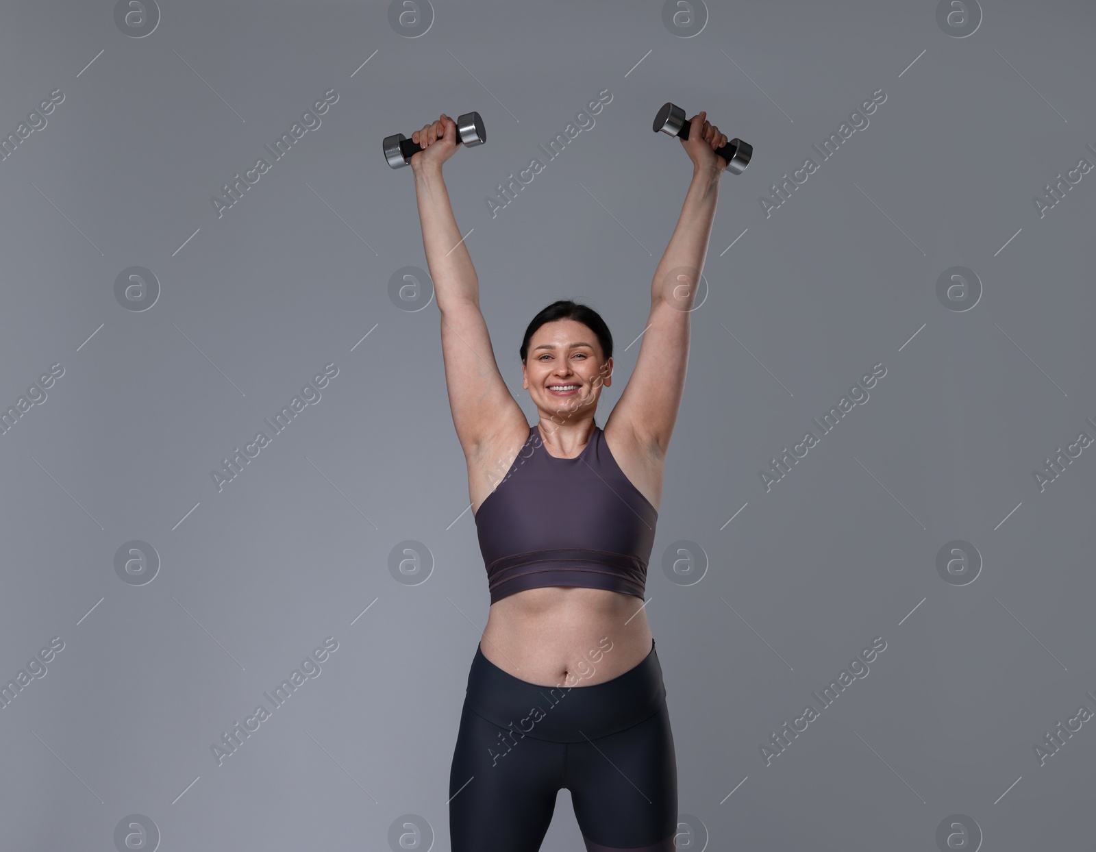 Photo of Plus size woman in gym clothes doing exercise with dumbbells on grey background