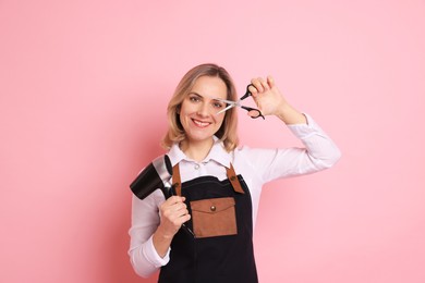 Smiling hairdresser with dryer and scissors on pink background