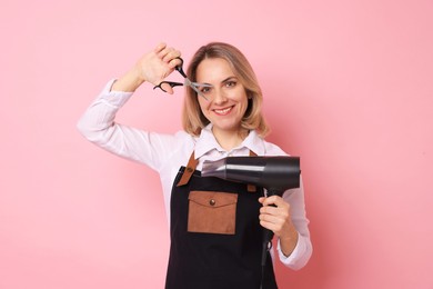 Smiling hairdresser with dryer and scissors on pink background