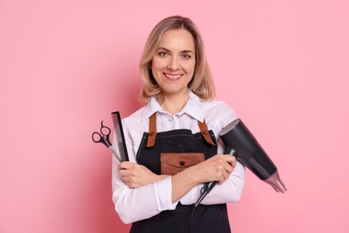 Smiling hairdresser with dryer, scissors and comb on pink background