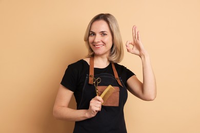 Smiling hairdresser with scissors and comb showing OK gesture on beige background