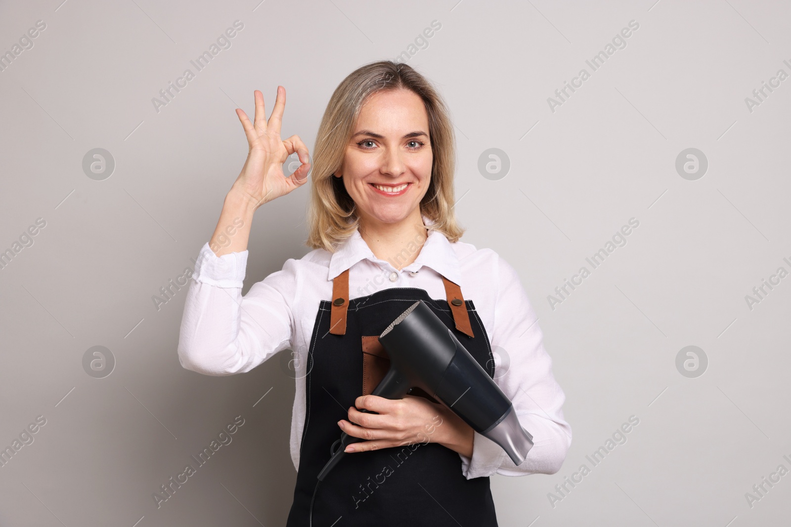 Photo of Smiling hairdresser with dryer showing OK gesture on gray background