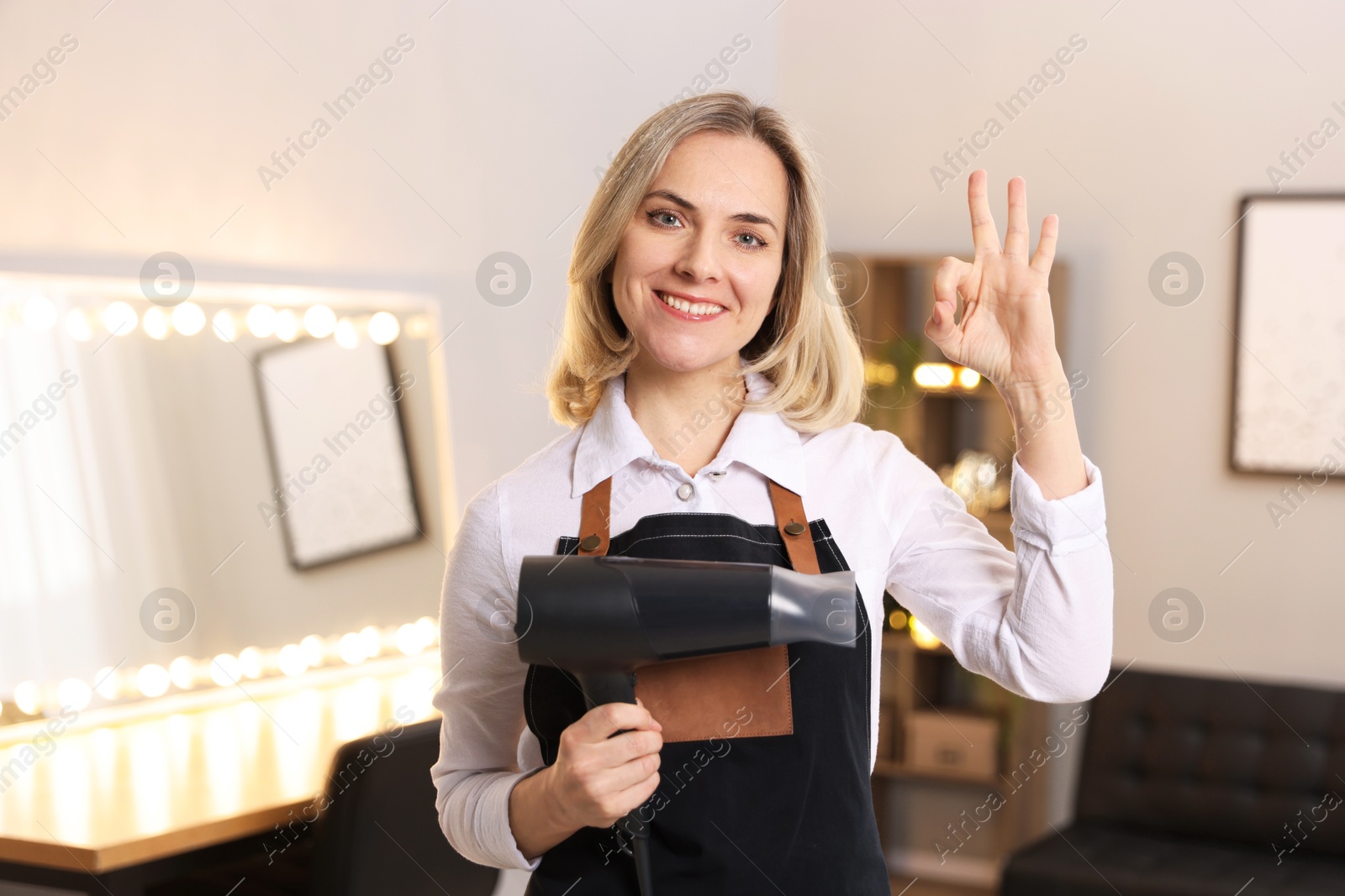 Photo of Smiling hairdresser with dryer showing OK gesture in beauty salon