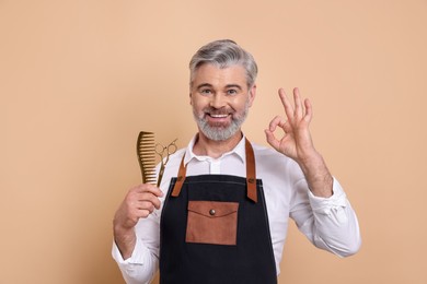 Photo of Smiling hairdresser with scissors and comb showing OK gesture on beige background