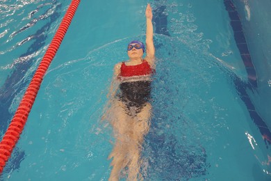 Photo of Sporty woman wearing cap and goggles swimming in pool indoors