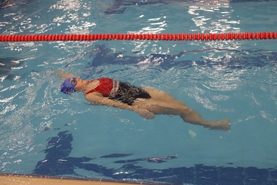 Sporty woman wearing cap and goggles swimming in pool indoors