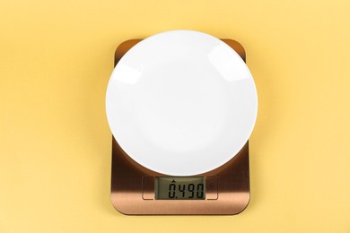 Photo of Electronic kitchen scale with plate on yellow background, top view