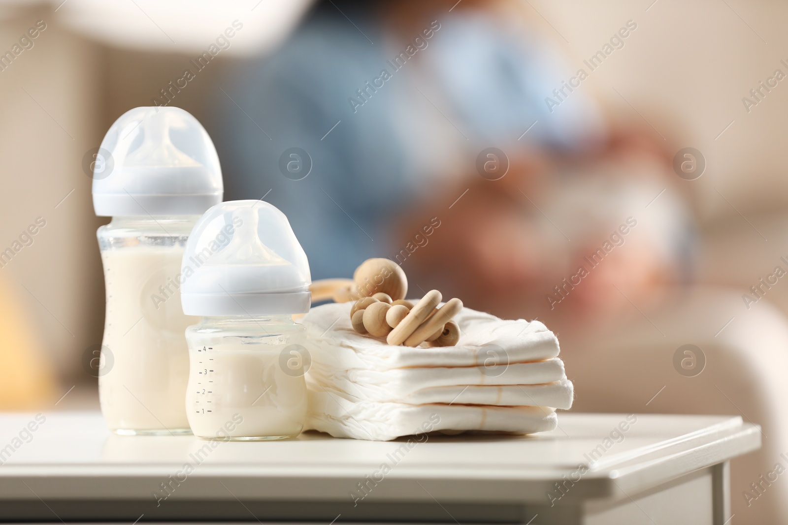 Photo of Mother holding her little baby indoors, focus on feeding bottles with milk, teether and diapers