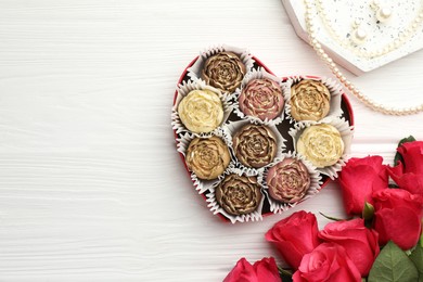 Photo of Flower shaped chocolate bonbons in box, red roses and pearl jewelry on white wooden table, flat lay. Space for text