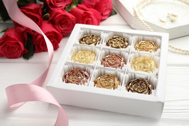 Photo of Flower shaped chocolate bonbons in box, red roses, pink ribbon and pearl jewelry on white wooden table, closeup