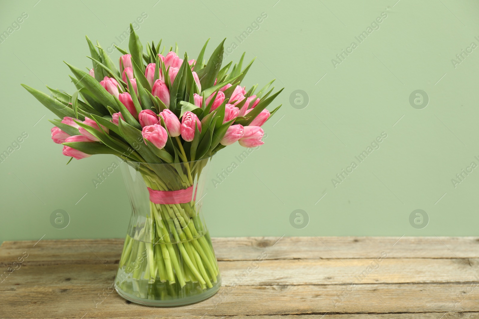 Photo of Bouquet of beautiful tulips in vase on wooden table against pale green background, space for text