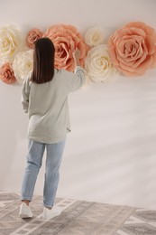 Photo of Woman decorating wall with beautiful paper flowers at home, back view