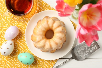 Photo of Delicious bundt cake, Easter eggs, tea and tulips on white wooden table, flat lay