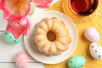 Photo of Delicious bundt cake, Easter eggs, tea and tulips on white wooden table, flat lay