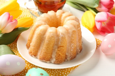 Photo of Delicious bundt cake, Easter eggs and tulips on white wooden table, closeup