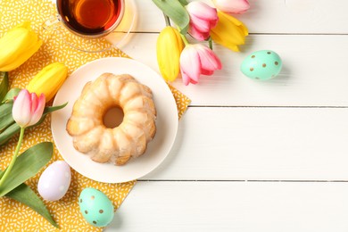 Photo of Delicious bundt cake, Easter eggs, tea and tulips on white wooden table, flat lay. Space for text