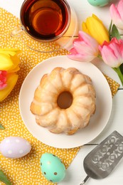 Photo of Delicious bundt cake, Easter eggs, tea and tulips on white wooden table, flat lay