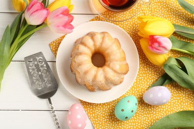 Photo of Delicious bundt cake, Easter eggs, tea and tulips on white wooden table, flat lay